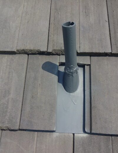 A gray vent pipe with sealant protrudes from a tiled roof, with one tile cut to fit around the base of the pipe.