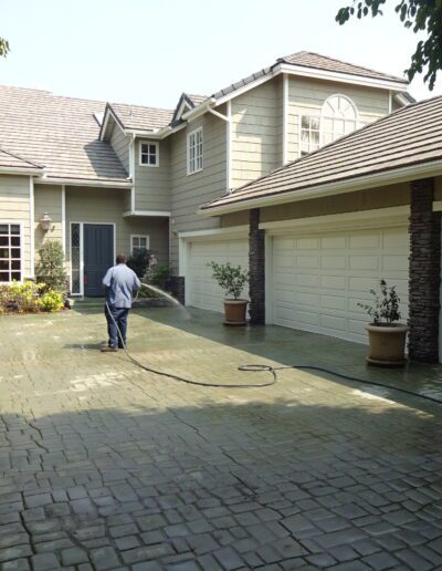 A person hoses down the driveway in front of a large house with three garage doors on a sunny day.